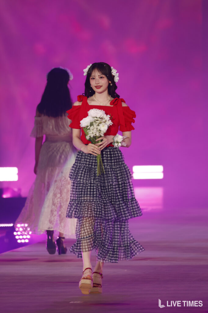 Young woman in a red off-shoulder top and tiered gingham skirt walks a runway, holding a bouquet of white flowers with flower crown on pink-lit stage.