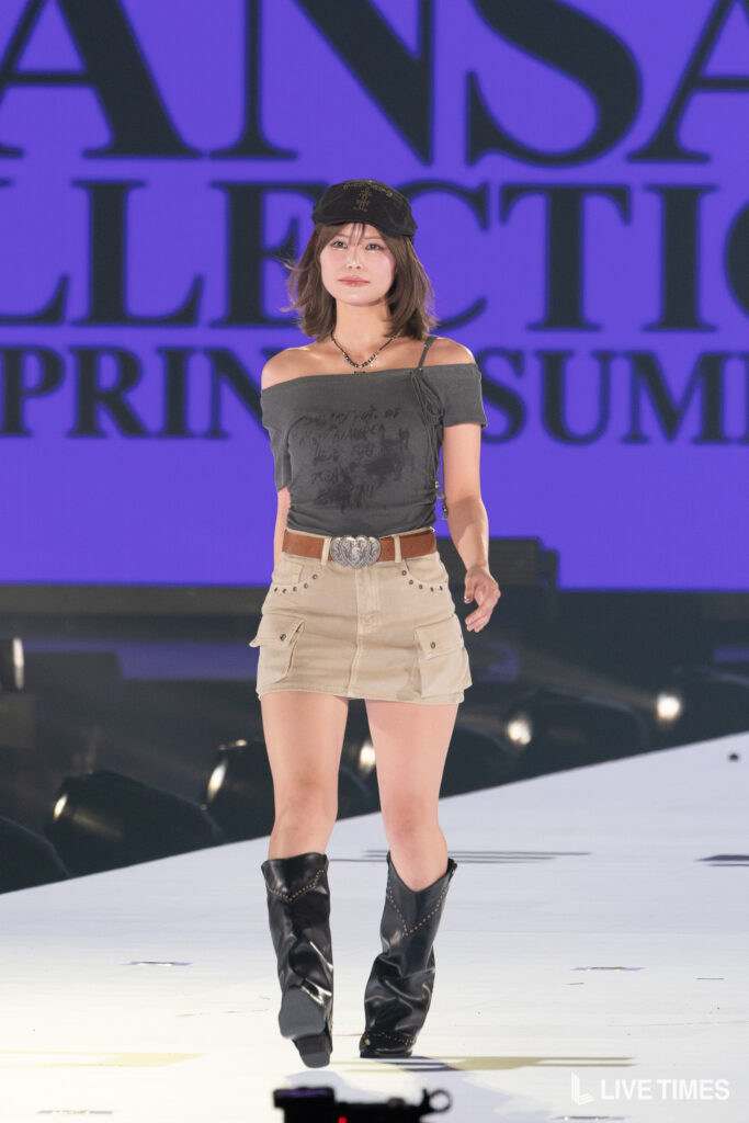 Woman model walking on a runway in a gray off-shoulder top, beige cargo skirt, and black boots; purple backdrop behind her.