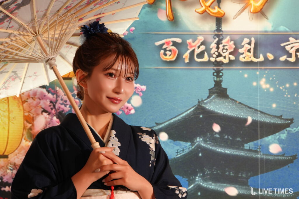 Woman in a dark blue kimono with floral patterns, holding a light wooden parasol, posed against a colorful festival backdrop with lanterns and Japanese script.