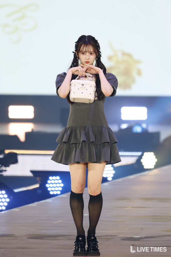 Young woman in a dark gray ruffled dress on stage, holding a small white handbag in front of her.