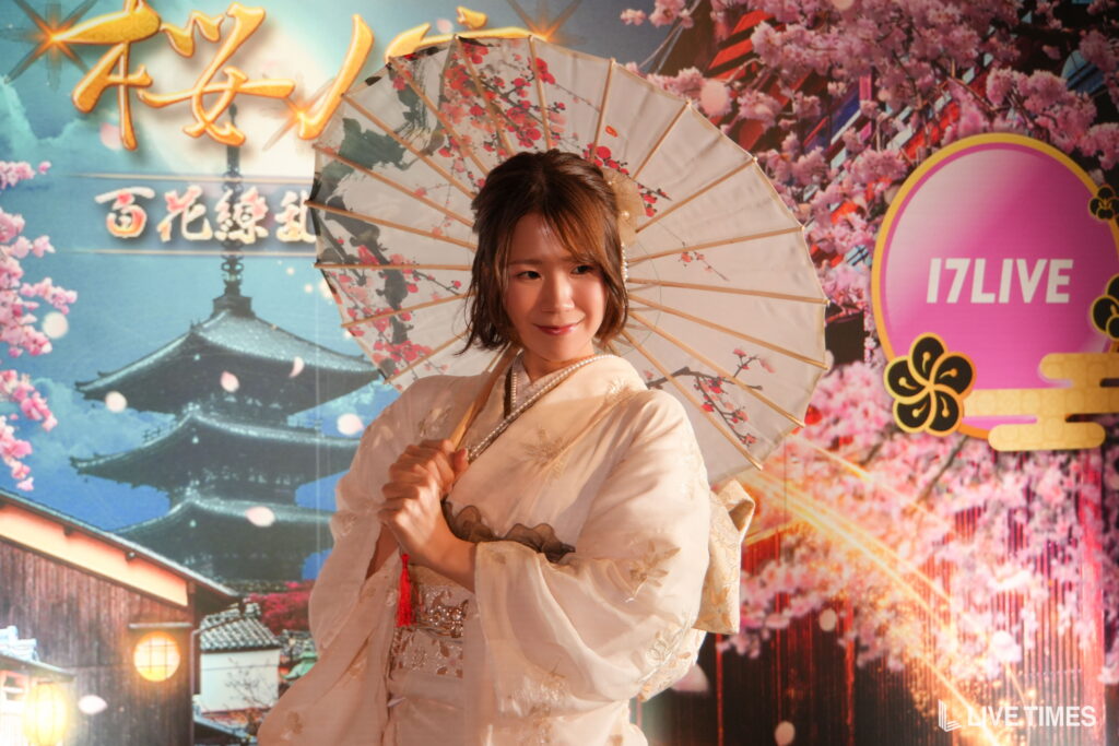 Woman in a pale kimono holding a floral parasol, smiling in front of a colorful festival backdrop.