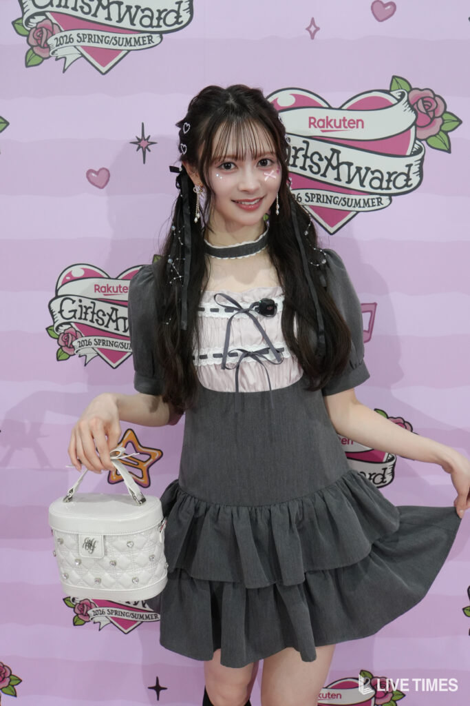 Young woman in a gray dress poses with a white quilted purse in front of a pink GirlsAward backdrop.