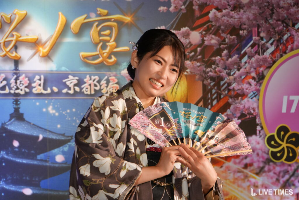 Smiling woman in a floral kimono holding colorful fans at a cherry-blossom themed event backdrop.