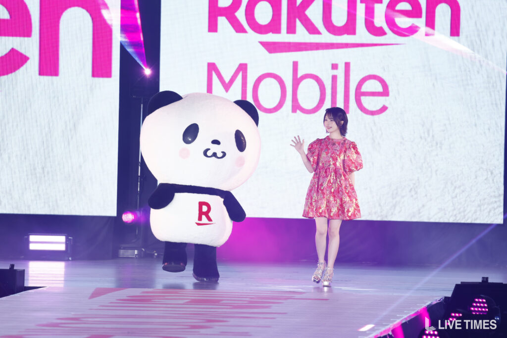 Panda mascot with red 'R' on belly posing with a woman in a pink dress on a stage at a Rakuten Mobile event.