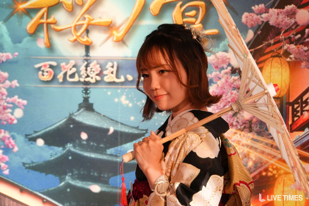Young woman in a floral kimono holding a white parasol at a colorful festival backdrop.