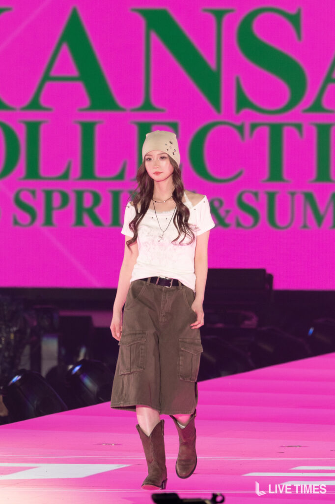 Female model walking a fashion runway on a pink stage with oversized green text backdrop; wearing a beige beanie, white tee, olive cargo skirt, and brown boots.