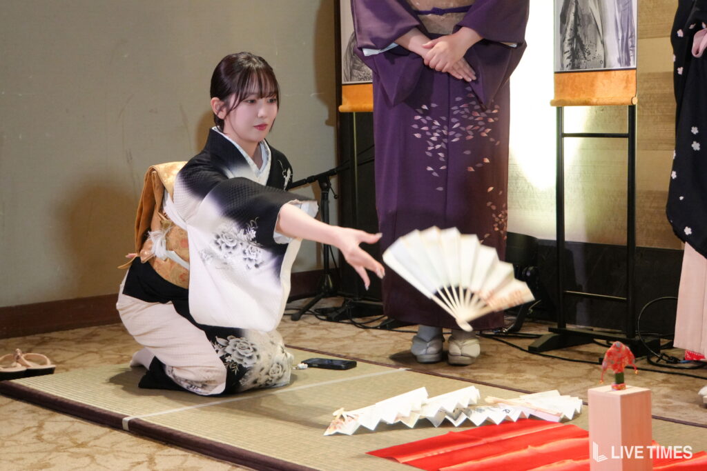 Woman in a white and black floral kimono kneels on a tatami mat, gracefully waving a white folding fan during a traditional performance.