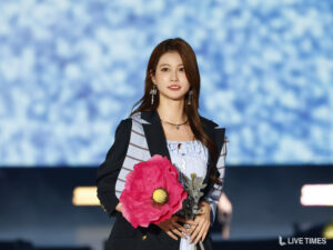 Woman on stage holding a large pink flower bouquet, wearing a dark blazer over a striped blouse with a blue blurred backdrop and statement earrings.