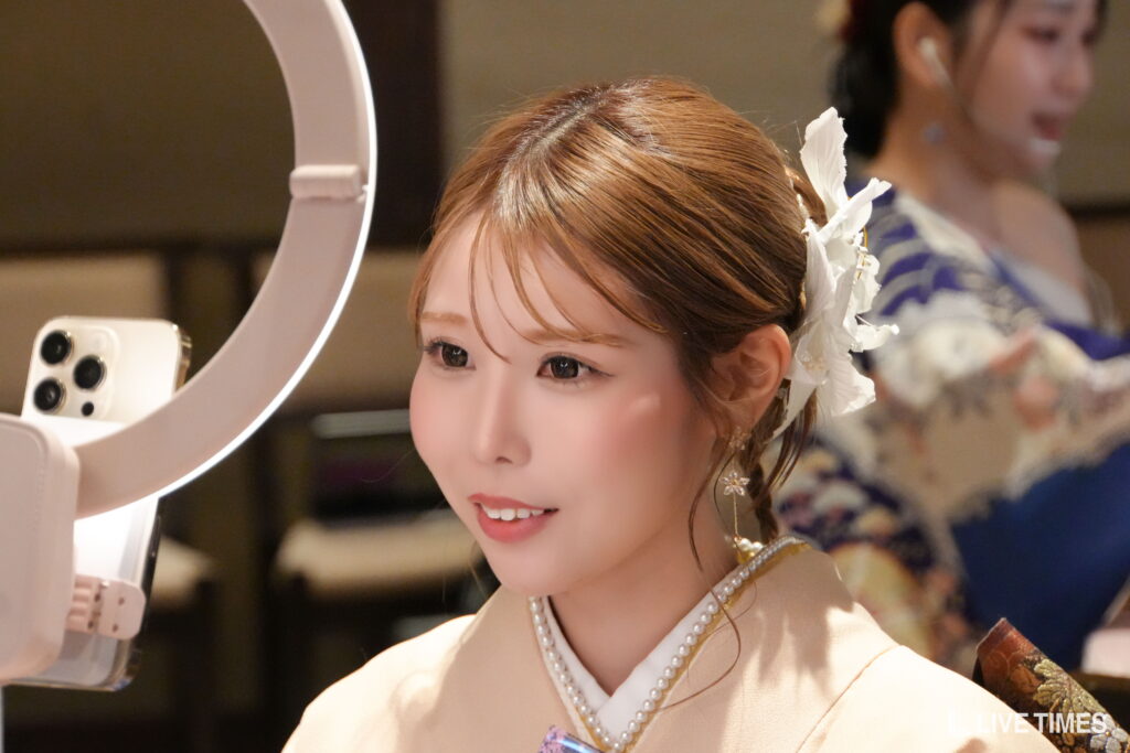 Close-up of a young woman with a floral hair accessory and pearl necklace, smiling at a ring light with a smartphone nearby, likely getting makeup or posing for a photo.