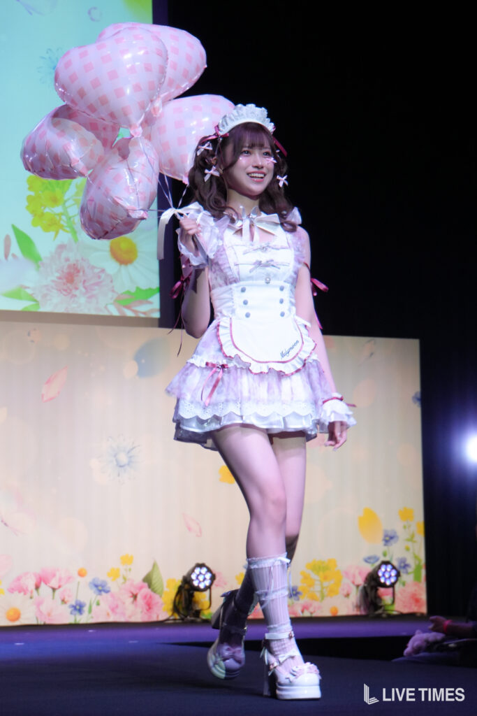 Young woman in a pastel pink maid outfit on stage, holding pink checkered heart-shaped balloons and smiling with a floral backdrop behind her.