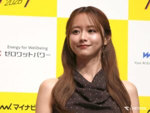 Close-up of a smiling woman with long brown hair in a black polka-dotted dress, standing before a yellow and white checkered backdrop at an event.