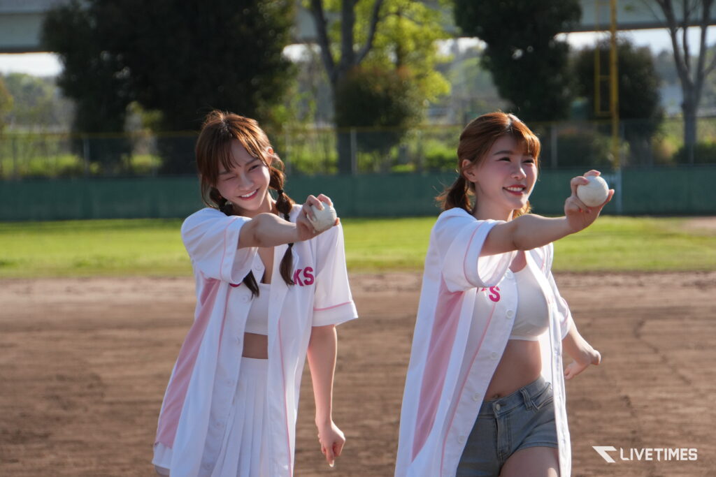 Two women in white baseball jackets with pink trim throwing baseballs on a sunny practice field, smiling at the camera.