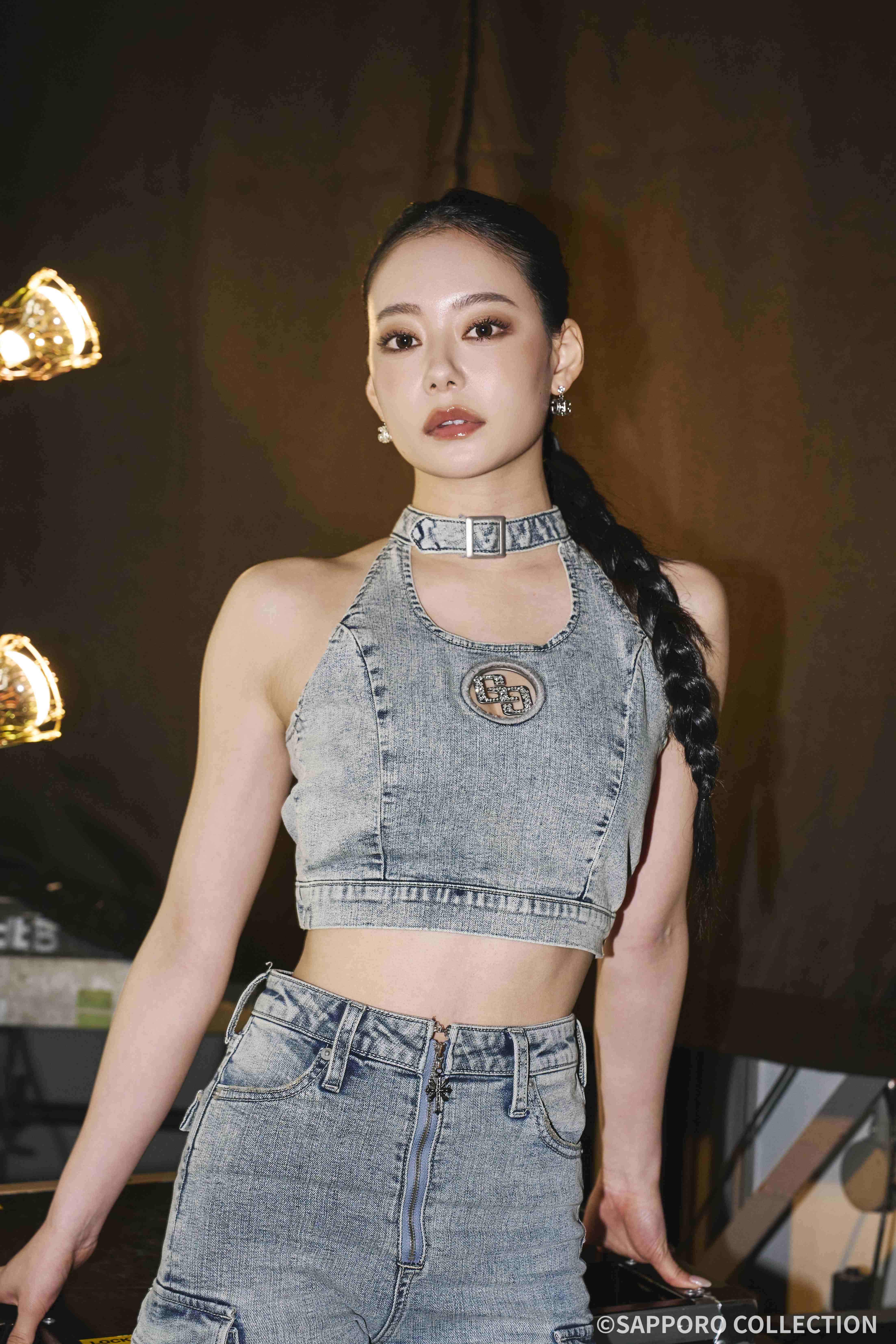 Woman in a sleeveless denim halter top with a braided ponytail, posing indoors against a dark backdrop.