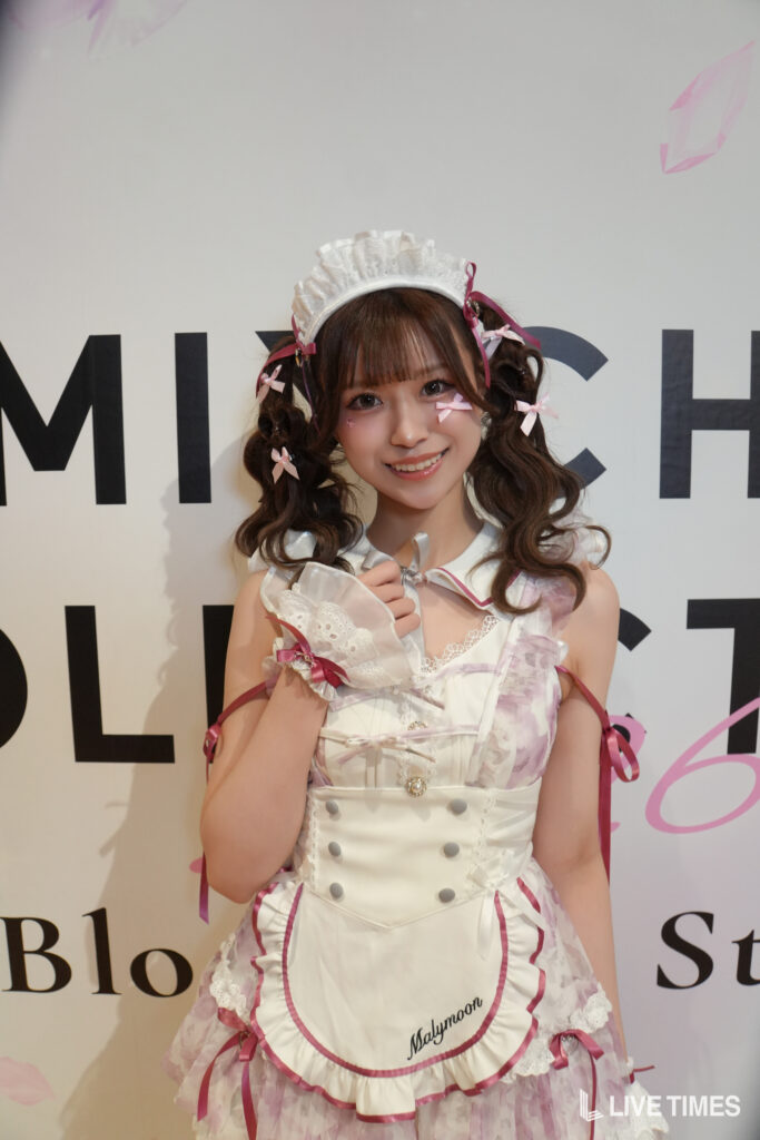 Young woman in a pastel pink-and-white maid cosplay, with a lace headband and pink ribbons, smiling at the camera against a bold lettered backdrop.