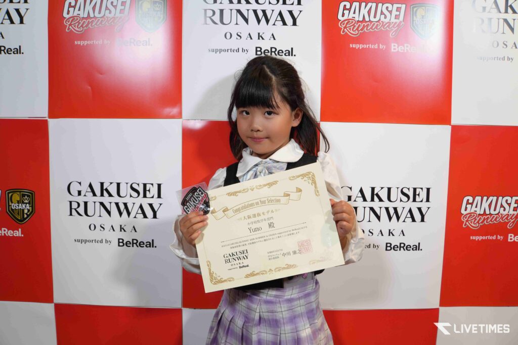 Young girl in a school uniform holding a certificate at a Gakusei Runway Osaka event backdrop.