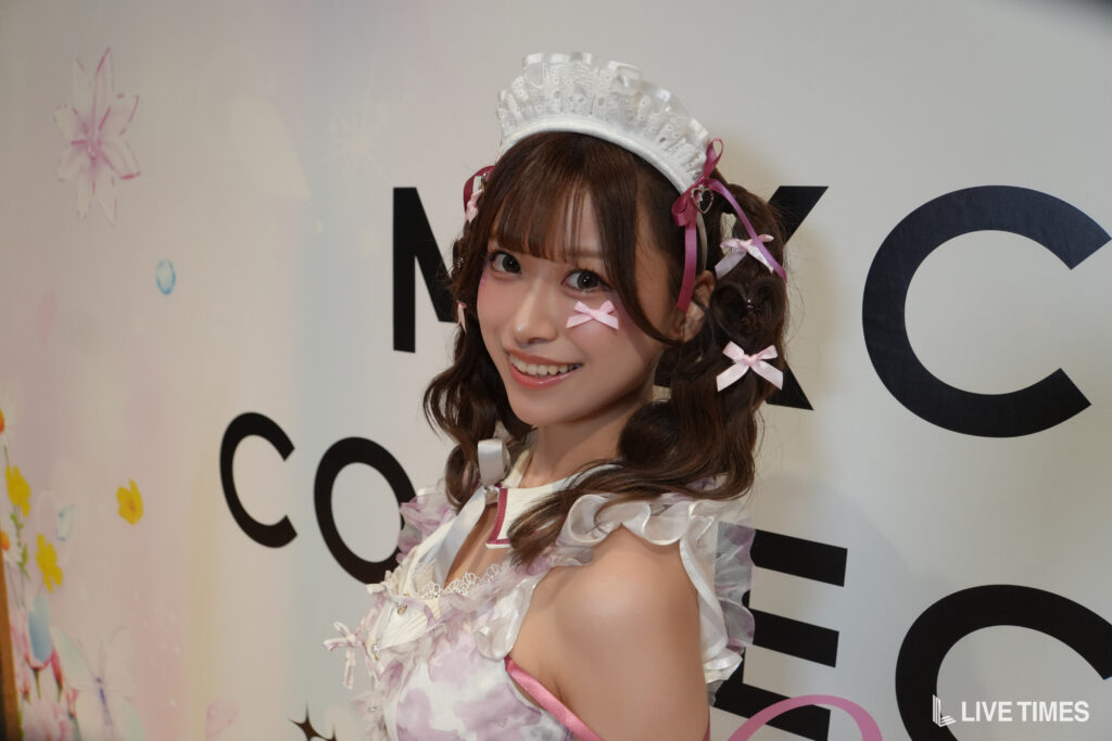 Young woman in a frilly white and pink maid cosplay, wearing a lace headpiece with pink ribbons, smiling at the camera in front of a banner.