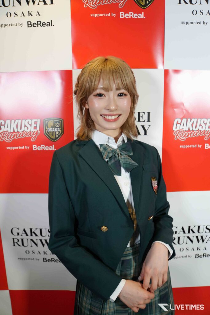Smiling young woman in a blazer and plaid skirt poses in front of a red-and-white backdrop with logos.