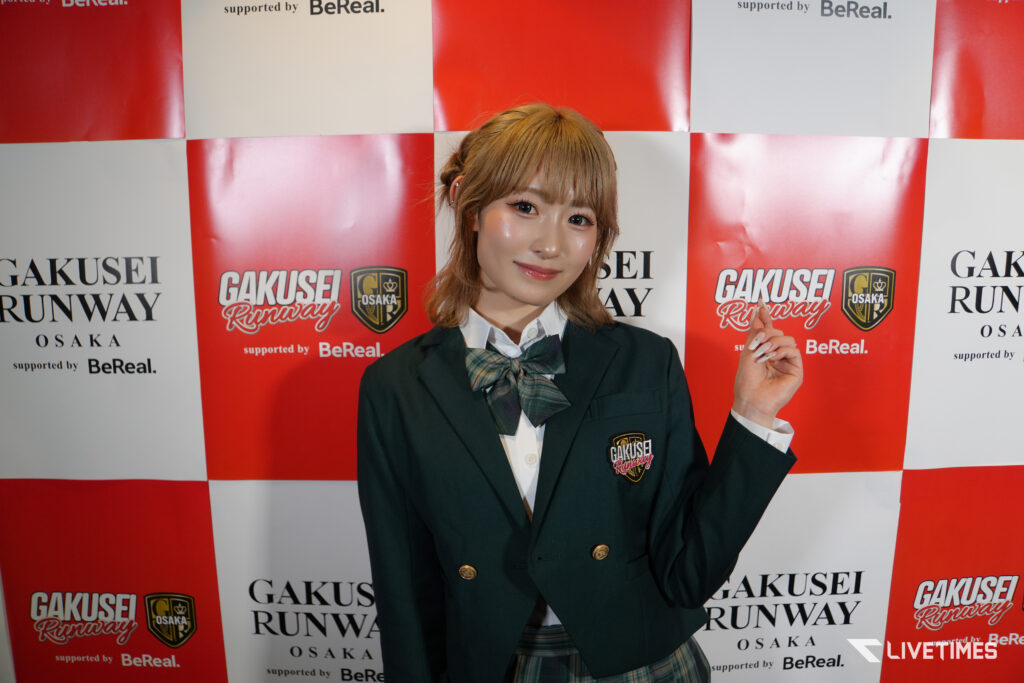 Young woman in a dark blazer and plaid tie posing at a Gakusei Runway Osaka backdrop.