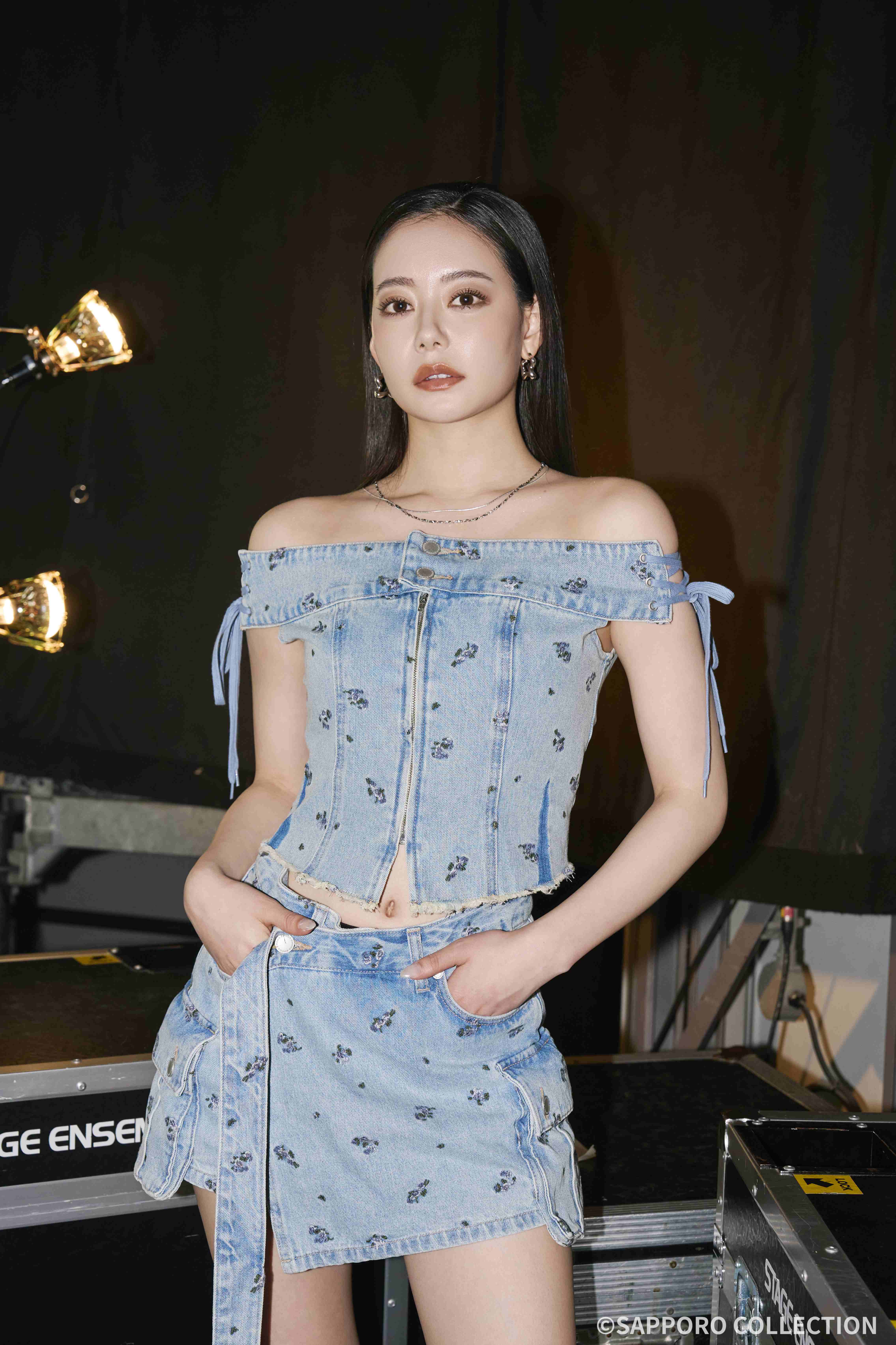 Young woman in an off-shoulder denim floral top and matching denim skirt poses backstage with stage lights and equipment in the background.