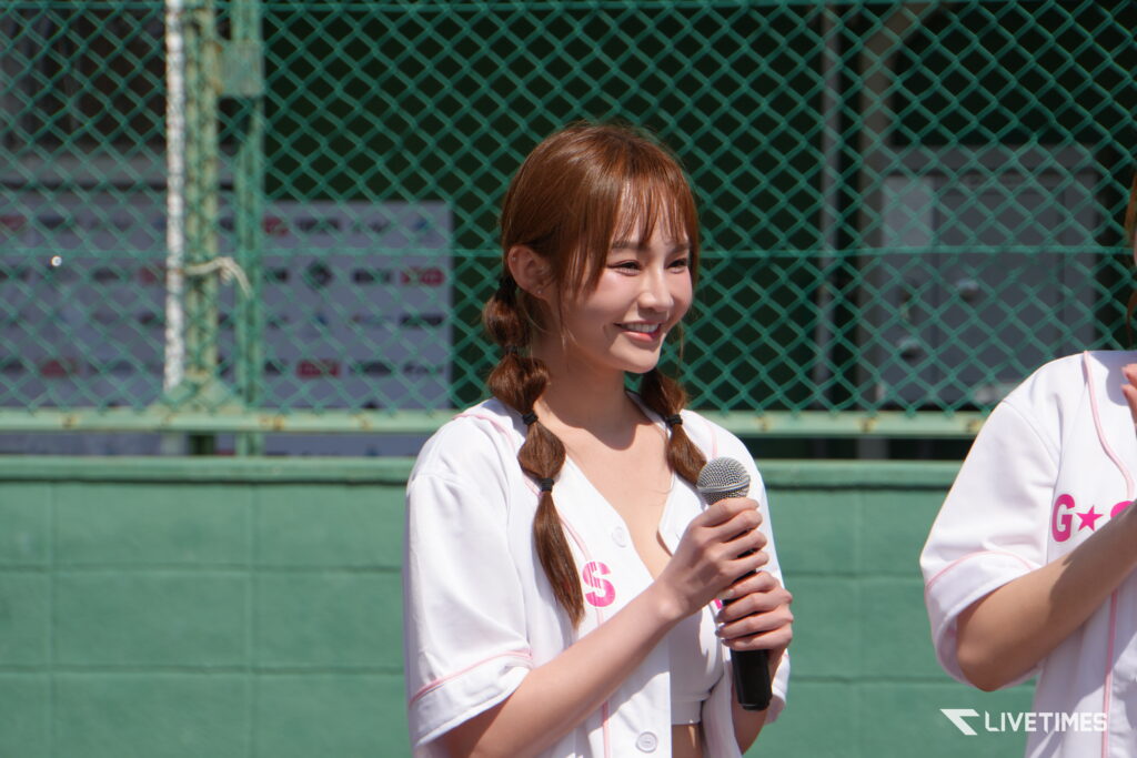 Young woman in a white outfit holding a microphone on a sunny outdoor stage with a green chain-link fence backdrop.