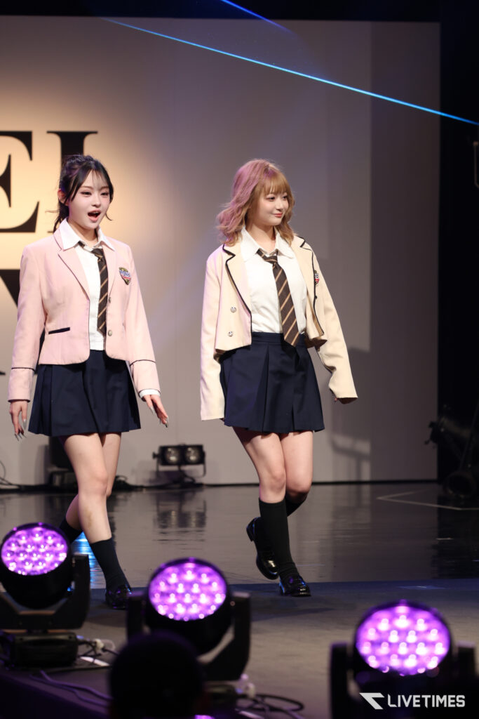 Two young performers in matching beige blazers, white shirts, striped ties, and navy skirts perform on a stage with purple lights.