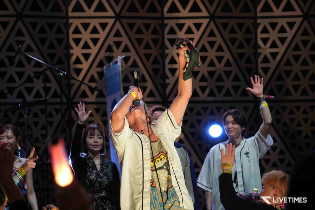 Live concert stage with a central performer raising a microphone while others cheer, geometric wall backdrop.