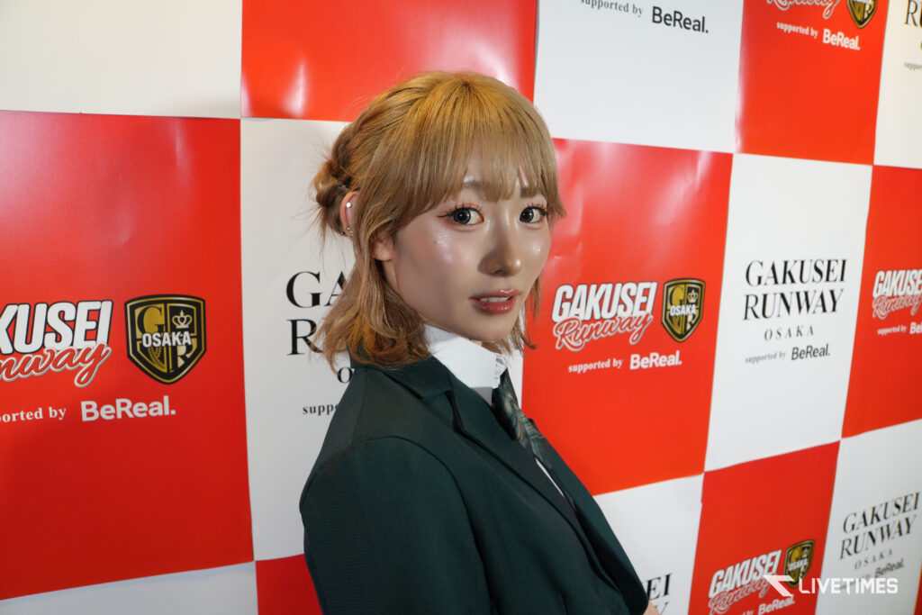 Portrait of a young woman in a dark blazer posing at a red-and-white sponsor backdrop featuring logos, including BeReal and Osaka.
