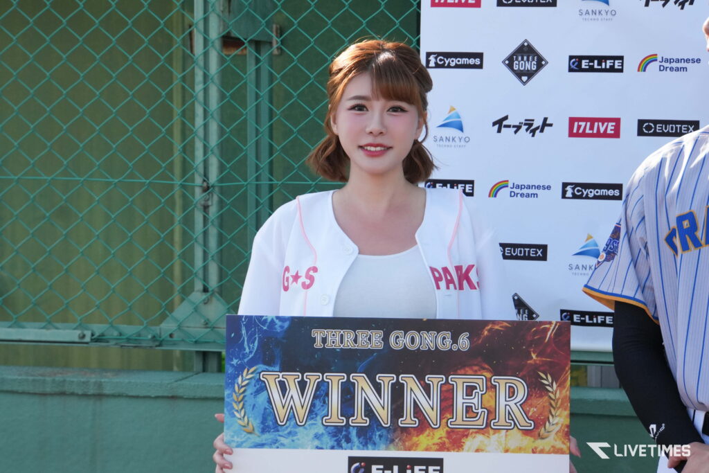Smiling woman with short brown hair in a white jacket holding a colorful WINNER sign at a sponsor-backed event backdrop.