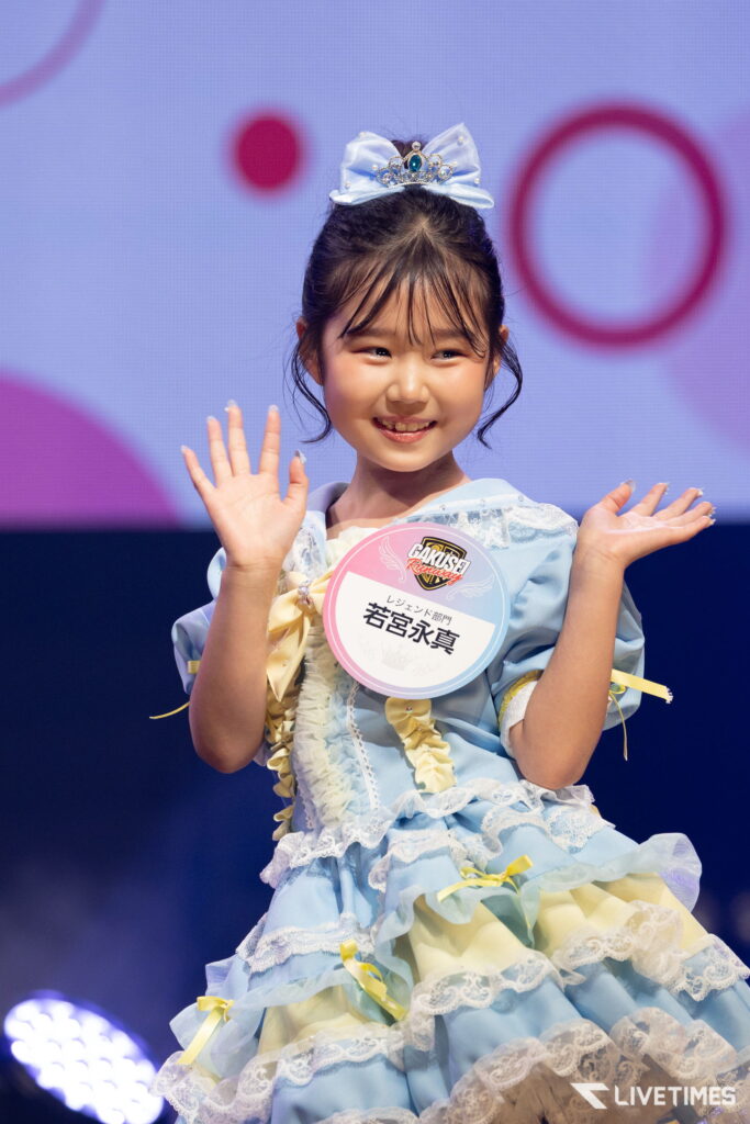 Young girl on stage in a blue frilly dress with a crown headband, waving both hands at the audience while wearing a round badge on her chest.