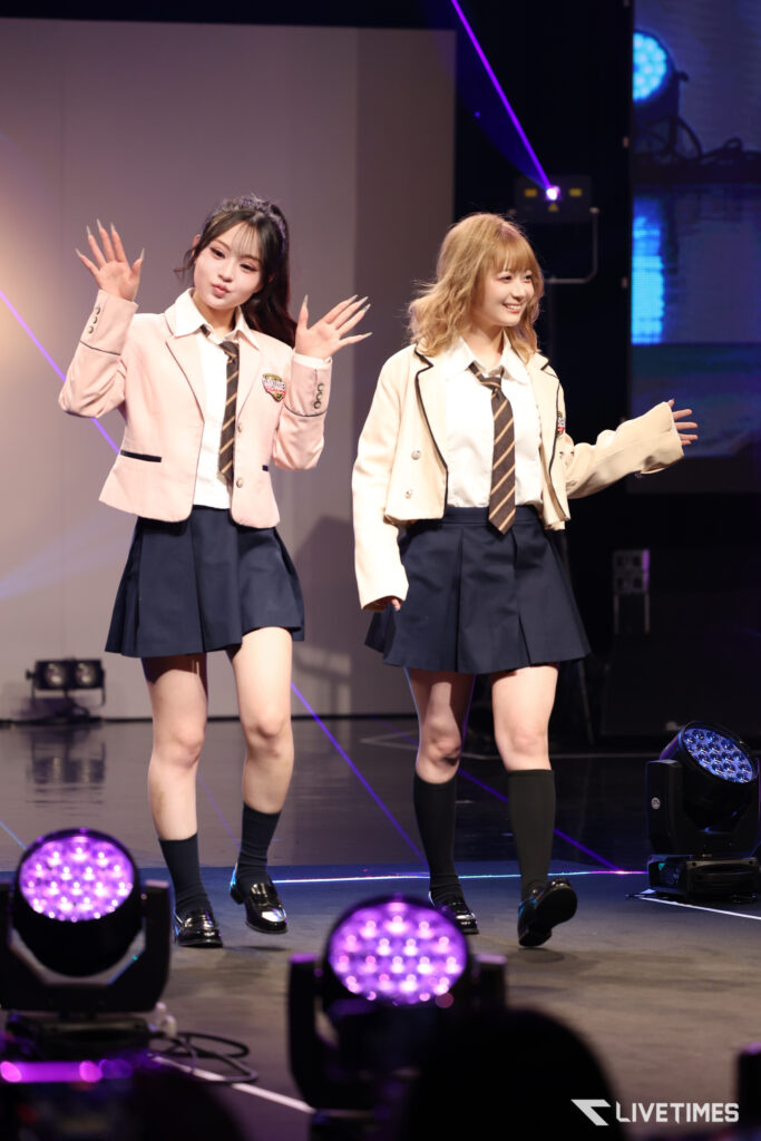 Two performers in beige blazers and navy skirts wave to the audience on stage.