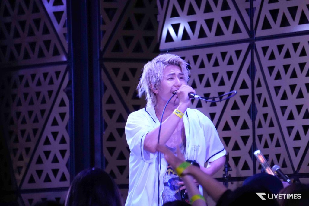 Male singer with blonde hair in a white shirt performs on stage under purple lighting, holding a microphone toward the audience.