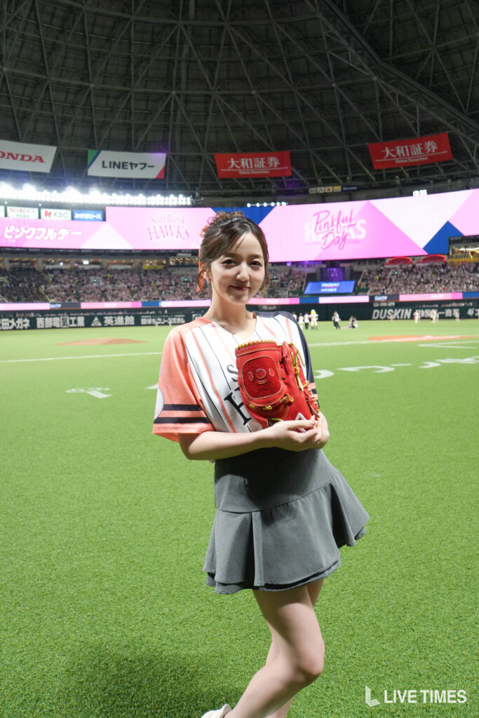 Smiling young woman in a pink-and-white striped shirt holding a red baseball glove on a baseball field with a large stadium and pink banners in the background.