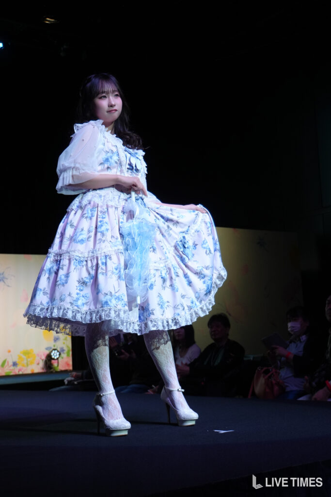 Young woman on stage in a light blue floral dress, lifting her skirt to reveal lace underskirt and white heels, spotlight on her.