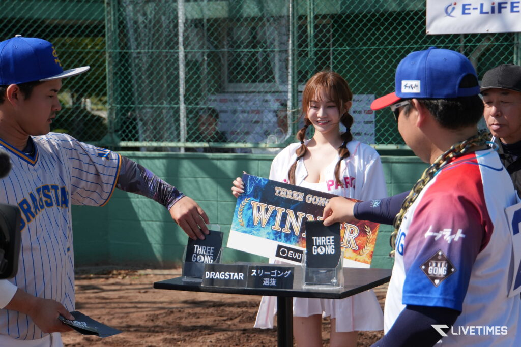 Young woman in a white outfit holds a colorful award board, flanked by players in blue-and-white jerseys at an outdoor ceremony.