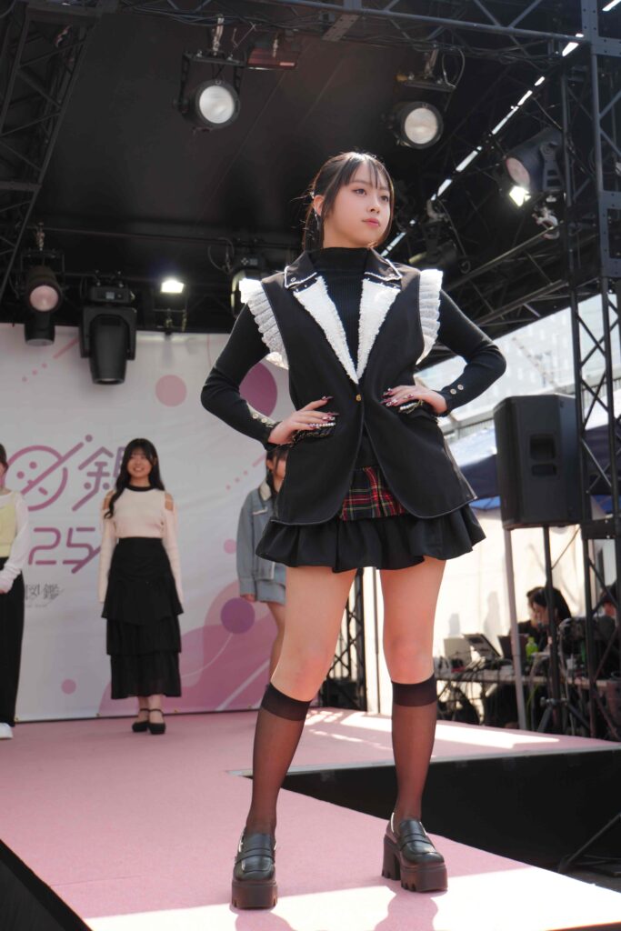 Model on stage wearing a black vest with white ruffled trim, plaid skirt, and chunky platform shoes; pink stage and lighting in background.