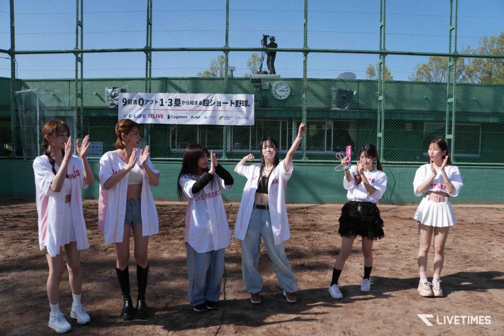 Girls in white jackets clap on a dirt baseball field, with a sponsor banner and a cameraman on a platform behind them.