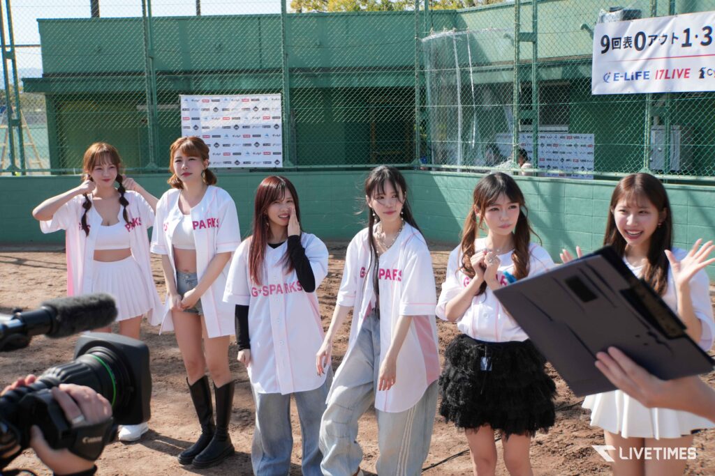 Group of young women in white uniforms posing for a camera outdoors, with a green fence and banners in the background.