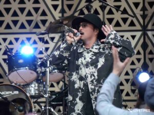 Male singer wearing a black hat and patterned shirt, holding a microphone on stage with drums in the background.