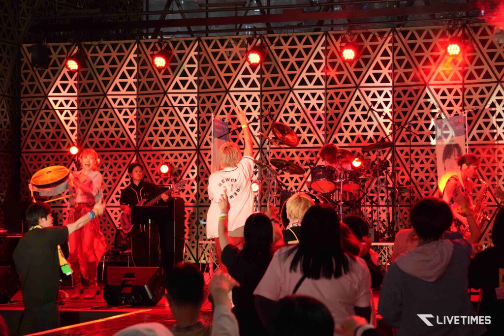 Band performing on a stage with guitarist and drummer under red spotlight, geometric backdrop behind them, and cheering audience in front.