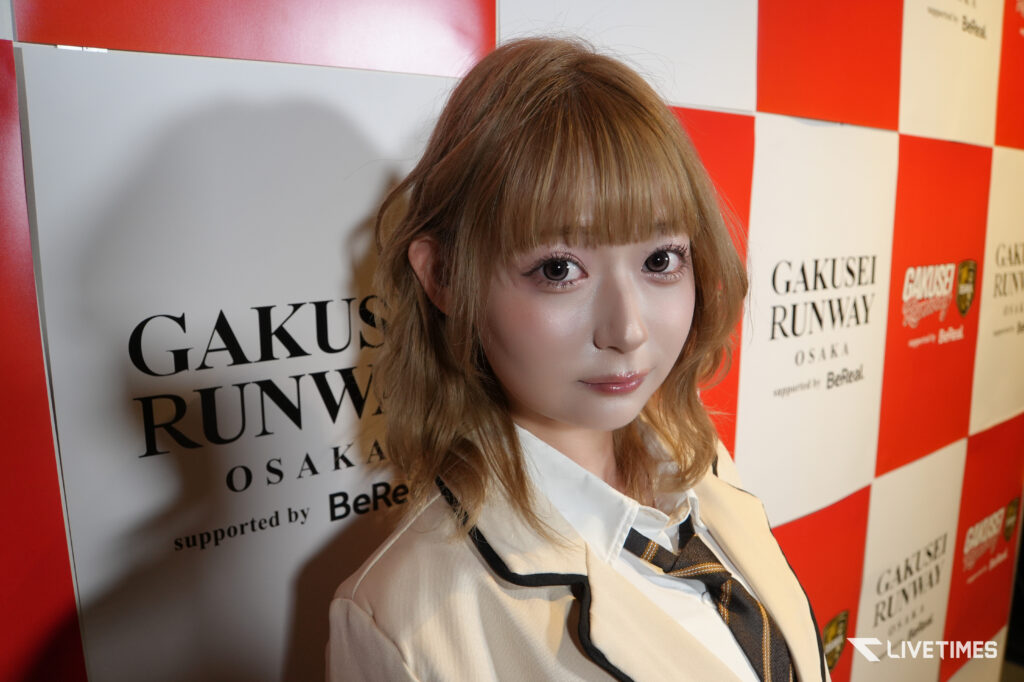Close-up of a young woman with blonde hair and makeup posing in front of a red-and-white checkerboard backdrop reading 'GAKUSEI RUNWAY OSAKA' (event wall).