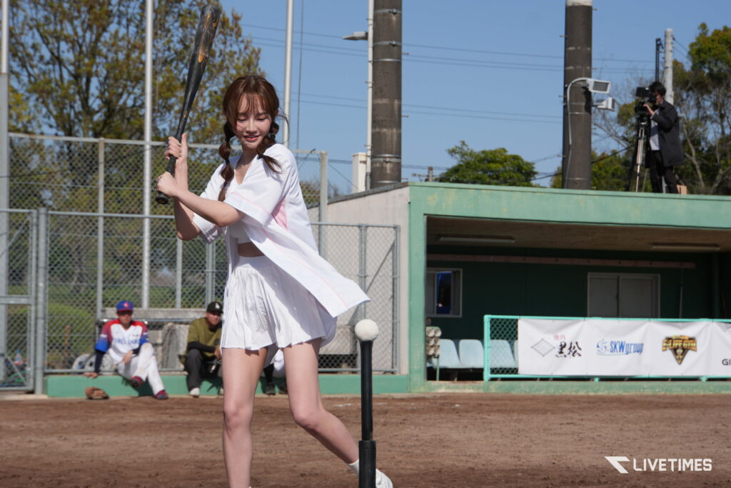 Young girl in a white baseball uniform swings a bat at a dirt field, with a fence and a cameraman in the background.