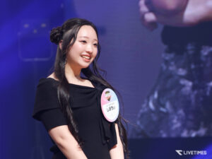 Smiling young woman with long black hair in a black dress on stage, wearing a round pink event badge on her chest.