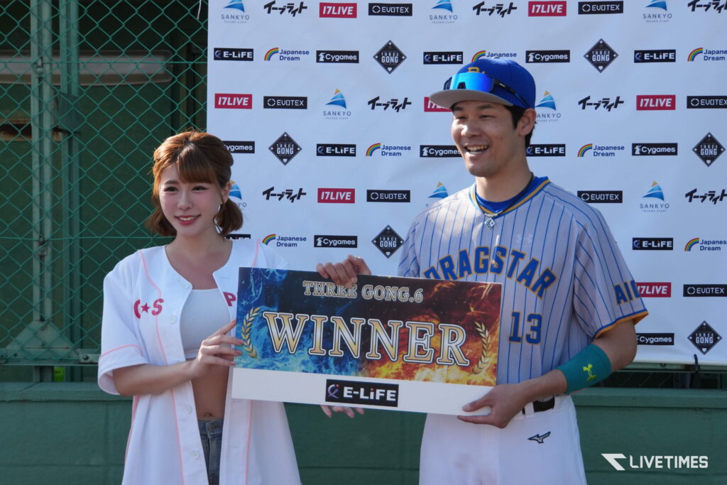 A smiling young woman in a white cardigan standing next to a baseball player in a striped uniform, both holding a colorful 'WINNER' sign in front of a sponsor backdrop.