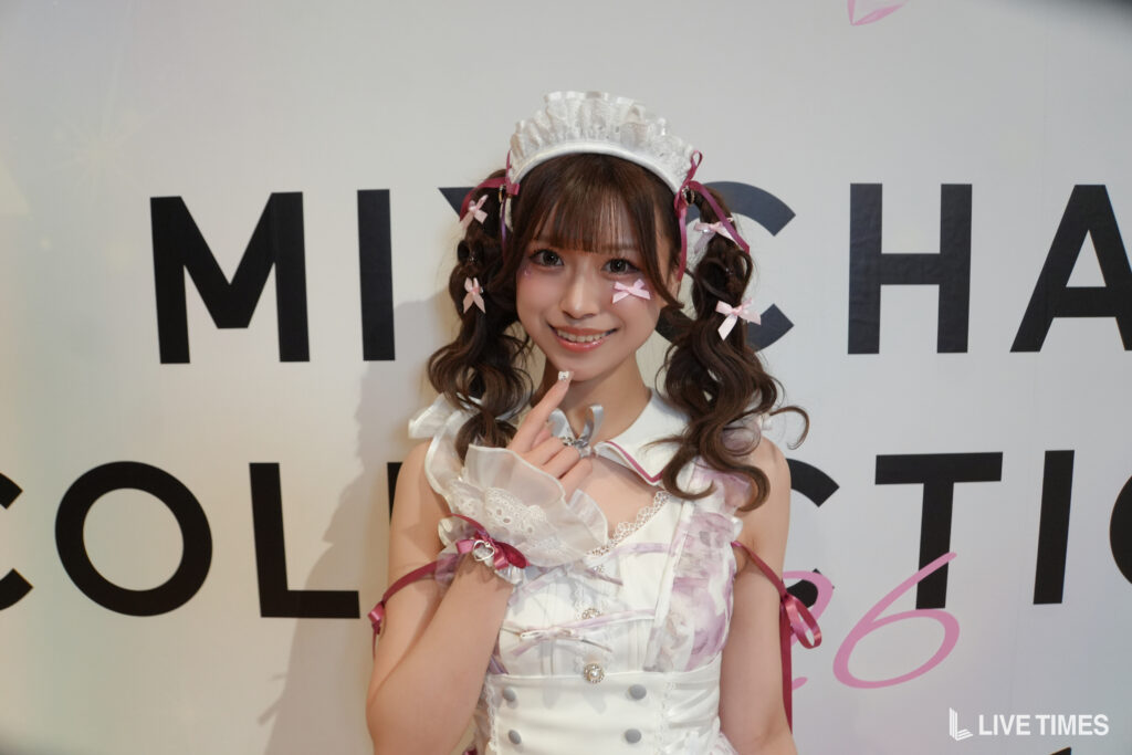 Young woman in a white and pink cosplay outfit with lace and bows, smiling at the camera against a wall with large black letters behind her.