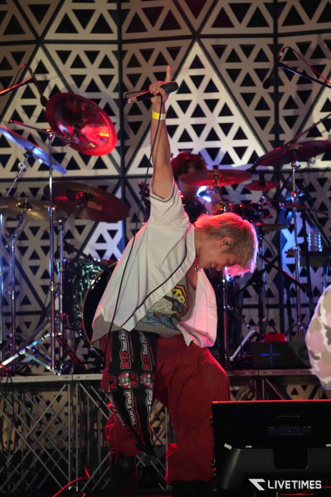 Singer on stage raises a microphone above their head; a drummer and geometric patterned backdrop are visible in the background.