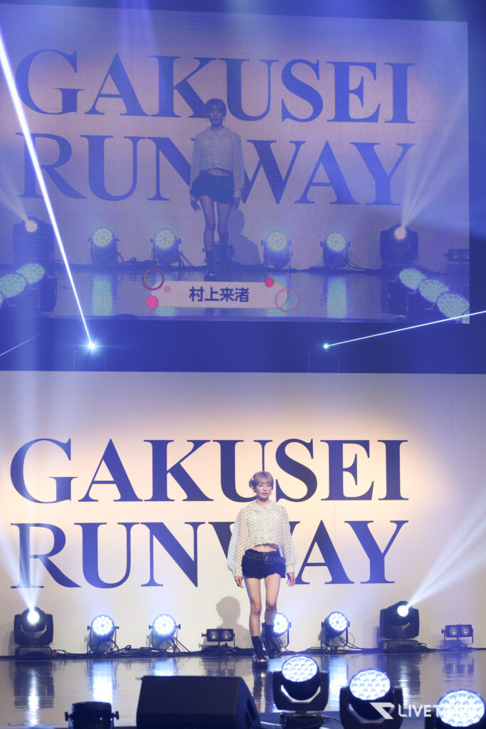 Model walks the runway in front of a large backdrop that reads 'GAKUSEI RUNWAY' with bright stage lights behind her.