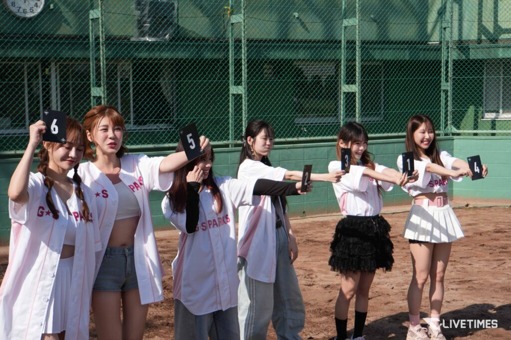 Group of women in white uniforms holding black numbered cards (6, 5, 3, 1, 4) on a dirt baseball field with a chain-link fence in the background.