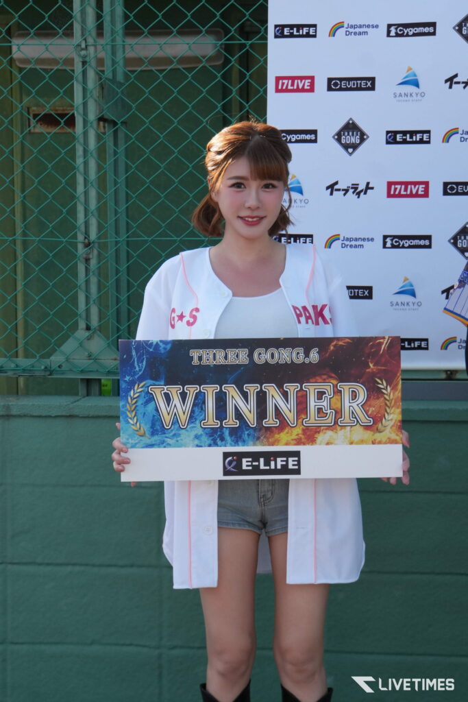 Young woman in a white jacket holds a large 'WINNER' sign at a sports event, smiles at the camera with sponsor logos behind her.
