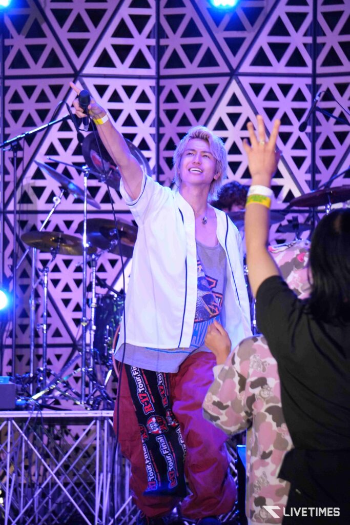 Performer on stage singing into a microphone with one arm raised, audience reaching toward him amid purple stage lighting and a geometric backdrop.