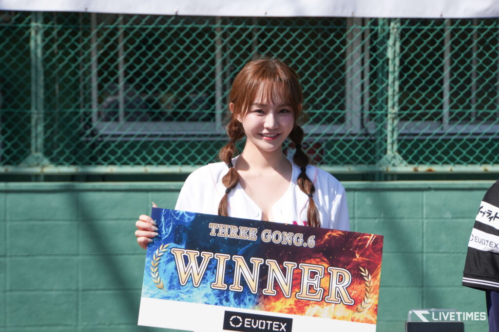 Smiling young woman holding a colorful banner that reads WINNER with EVOTEX logos, standing in front of a green chain-link fence.